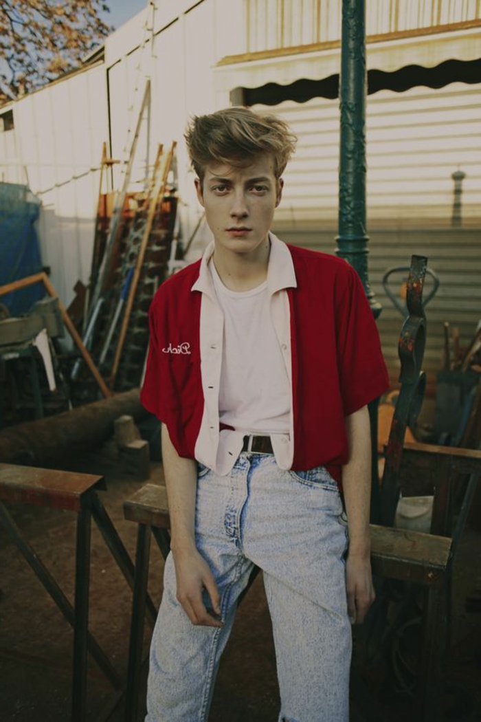 80's fashion for men, blonde boy with gelled up messy hair, wearing white t-shirt and red jacket with cropped sleeves, light blue acid wash jeans, messy yard in background