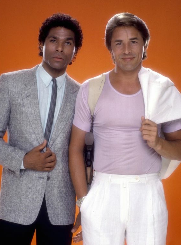 80s clothing, Miami Vice tv show style, African-American man in suit with thin tie, grey blazer and black trousers with white shirt, Caucasian man in pink t shirt with white trousers, holding white blazer over his shoulder