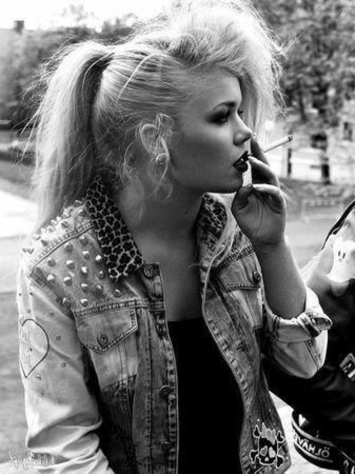 80s fashion, punk girl in black and white photo, heavy make up and smoking a cigarette, studded denim jacket with animal print collar and black top, park and trees in the background