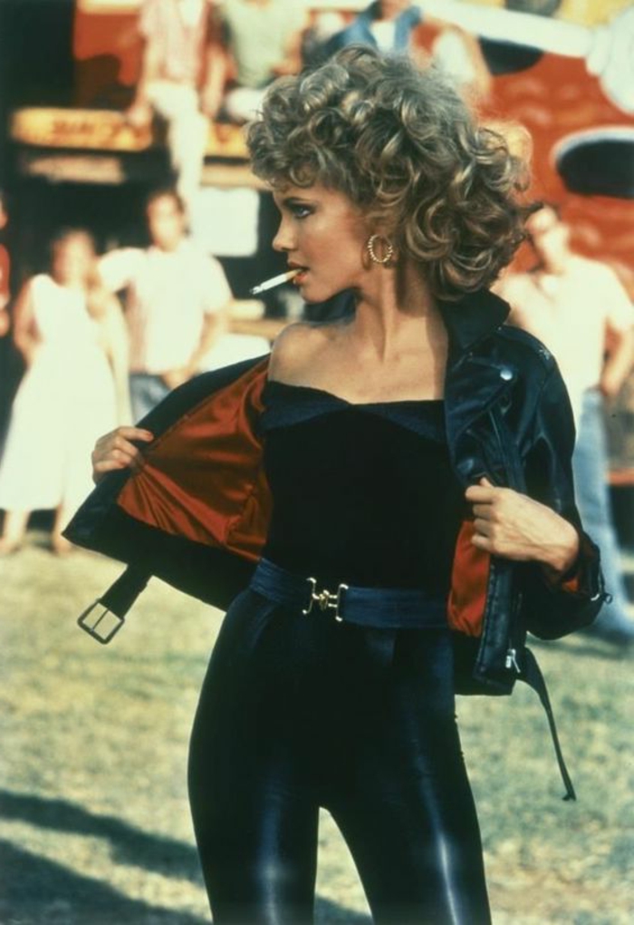 80s fashion trends, Grease, Olivia Newton-John, big curly blonde hair perm, black top and shiny black leggings, black leather jacket with red lining, wide belt and a cigarette