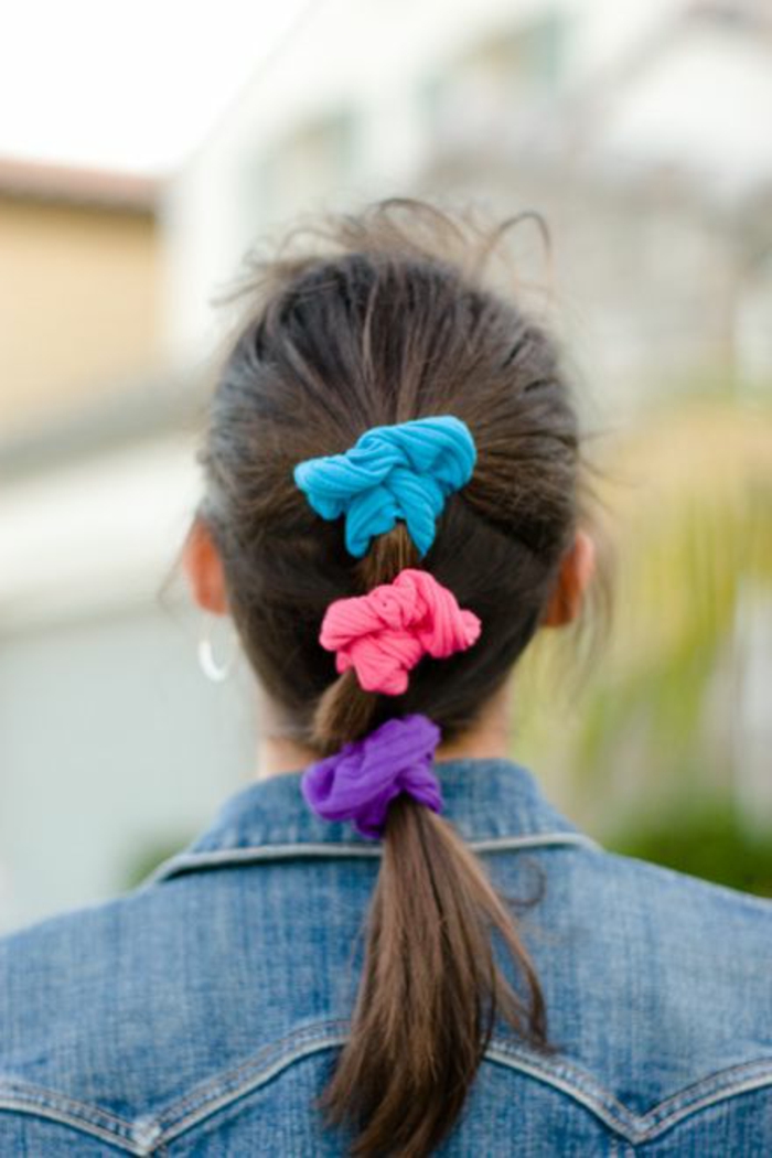 80s clothing, woman facing backwards with brown mid-length hair tied in ponytail, three bright neon scrunchies in blue pink and purple, jeans jacket and smudged background