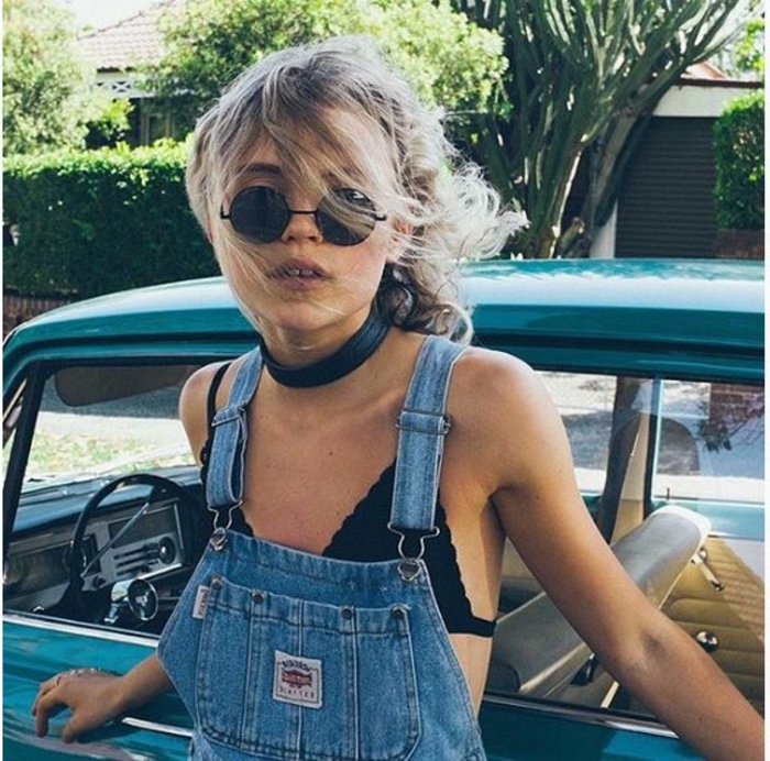 blonde girl with messy hair black choker and dark round sunglasses, wearing blue overalls over black bra, leaning on a retro blue car