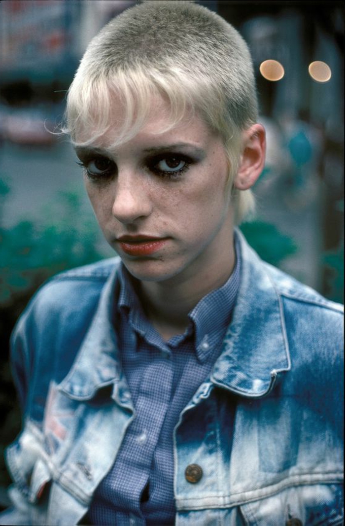 girl with mullet, blonde very short hair with longish bangs, black eye make up and red lipstick, wearing stone washed denim jacket and blue shirt