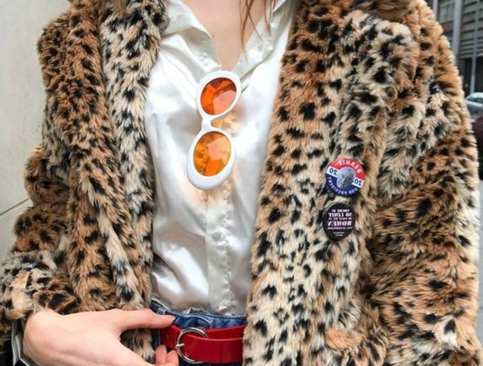 close up of a woman's torso, wearing white silky shirt and faux fur coat with animal print, pins and white sunglasses with orange frames, hand on red belt