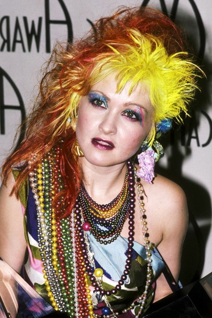 cyndi lauper with a messy punk hairstyle is neon yellow and orange, blue and pink glittering eye shadow, lots of necklaces with colorful beads, dark pink lipstick and shiny top