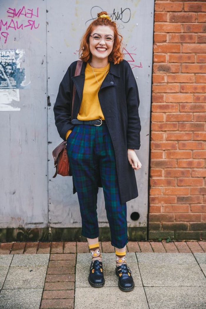 smiling woman with curly ginger hair and a top knot, navy blue coat over mustard yellow jumper, cropped blue and green tartan trousers, brown bag and owl socks