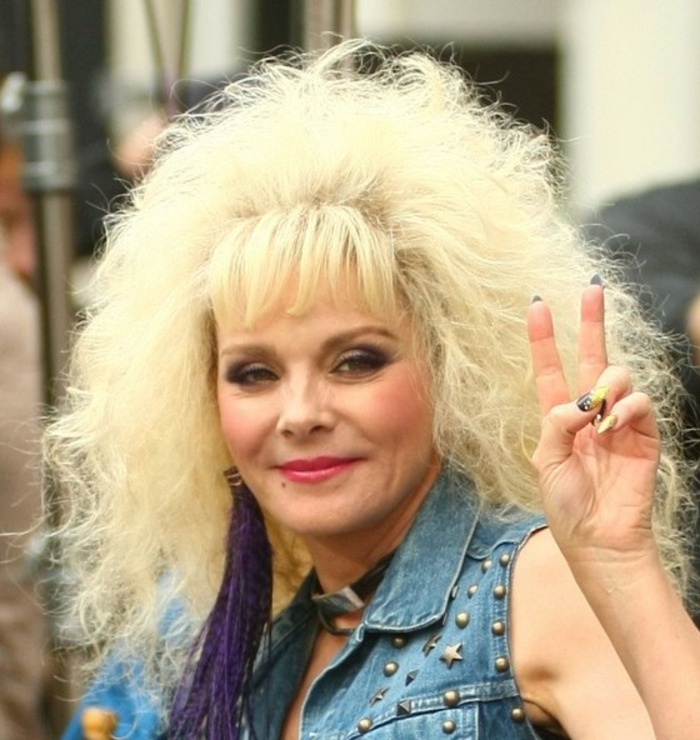 80s fashion trends, Kim Cattrall with big bleached feathery hair perm, star studded denim gilet, choker and big purple earring with tassels, holding a peace sign