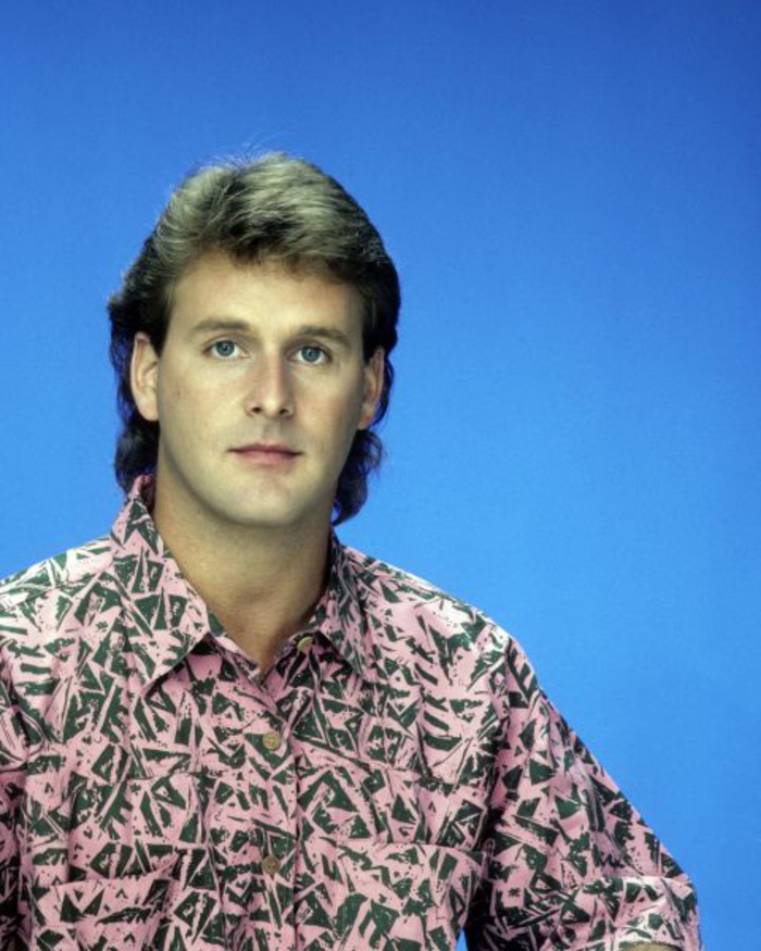 80's fashion for men, man with pale pink shirt, scribbles or geometric print, light brown mullet, plain blue background