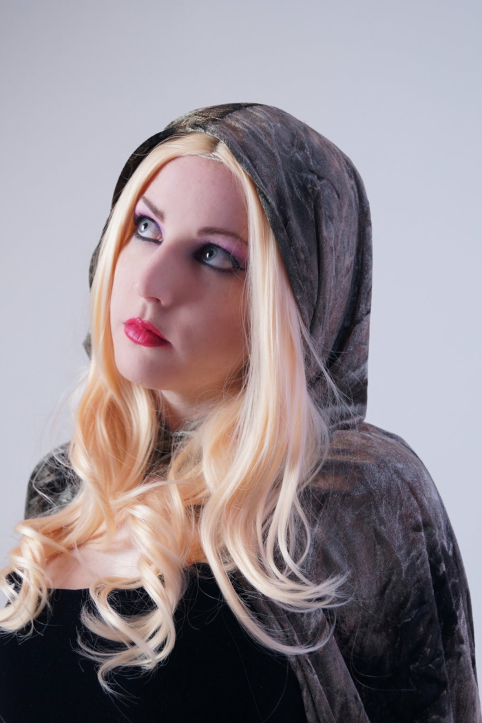 woman with red lipstick, black eye-make-up and blonde wig, wearing a black top, and dark grey velvet cape and hood