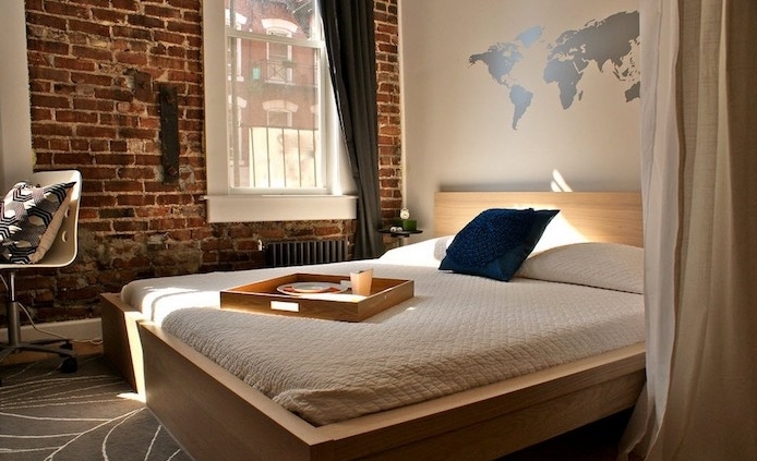 brick wall inside a sunlit room, other visible wall is white, with a silver world map wall art décor, simple bed with wooden food tray