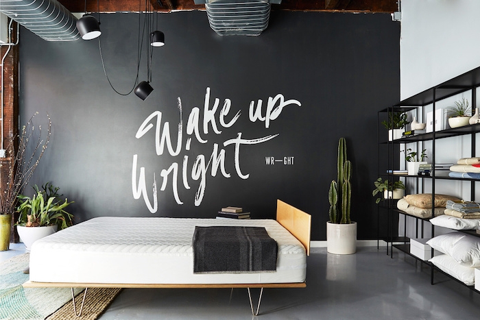 black wall with large bold white writing, inside industrial-style room, smooth pale gray floor, hanging lights and pipes on the ceiling, master bedroom ideas, black metal shelves