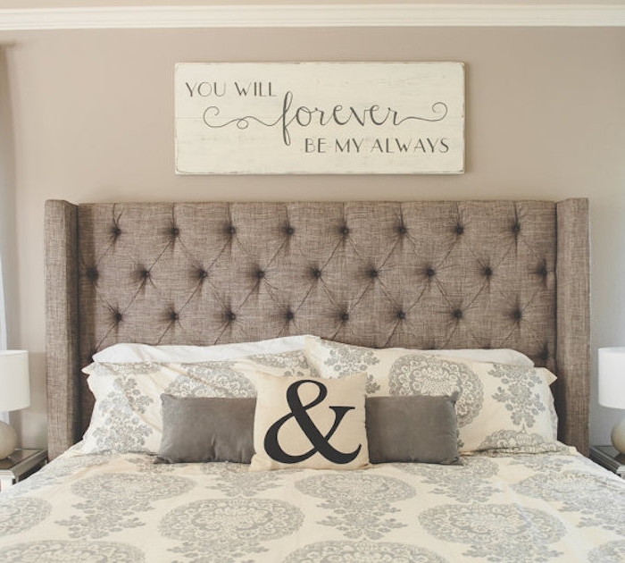 romantic message spelled in gray, on a cream-colored wooden board, hanging over a double bed, with beige soft headboard, and cream and gray bedding