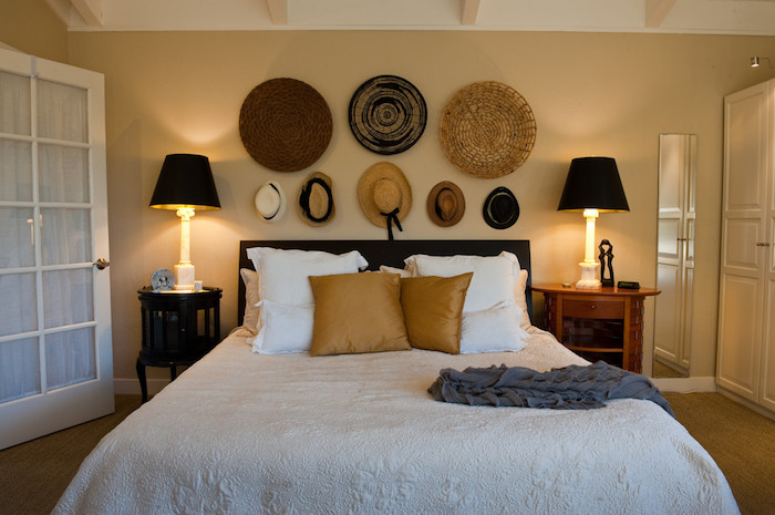 hats and round mats in different sizes and colors, made from straw, decorating a pale yellow wall, near double bed, with two bedside tables, and matching lamps