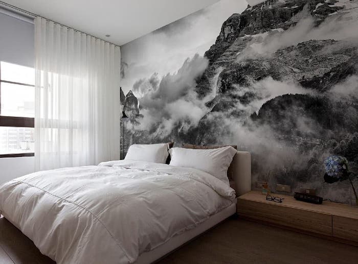 large wall art, grayscale photo wallpaper depicting cloudy mountains, near bed with fluffy white duvet, and two matching pillows, wooden cupboard and window