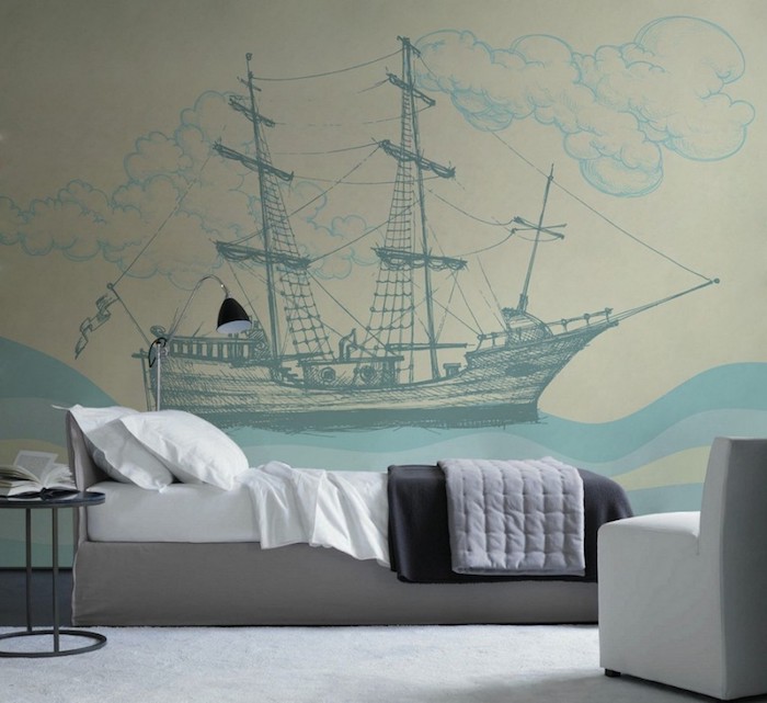 drawing of an antique ship, waves and clouds, done in different shades of blue, on a wall near a small, gray and white single bed, bedroom decorating ideas