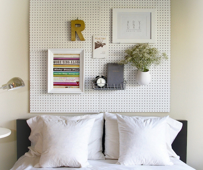 small flower vase with white flowers, two framed images, wire shelf with book and alarm clock, attached to a white board, hanging over a double bed, bedroom design