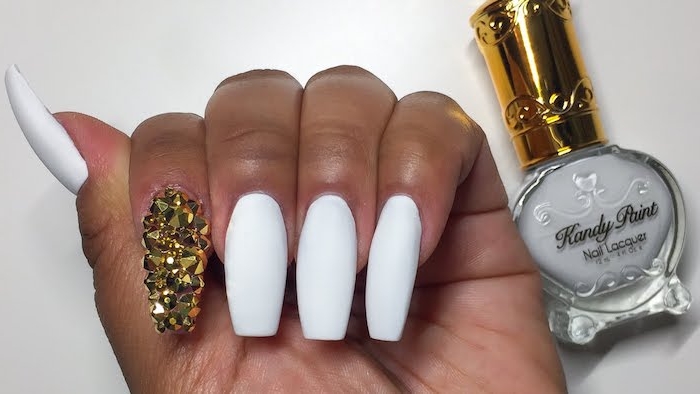 coffin acrylic nails, painted in white matte nail polish, the index finger nail, is covered in gold rhinestones, bottle of white nail polish nearby