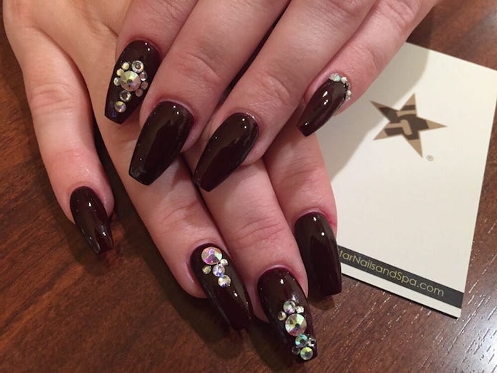 very dark cherry red nail polish, decorating the eight visible nails of two hands, ballerina nail shape, with rhinestones in different sizes
