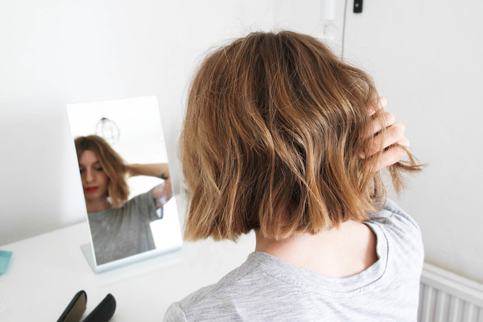 young woman looking at her reflection in a mirror, placed on a white desk, near some straightening irons, short haircuts for fine hair, light chestnut brown wavy bob