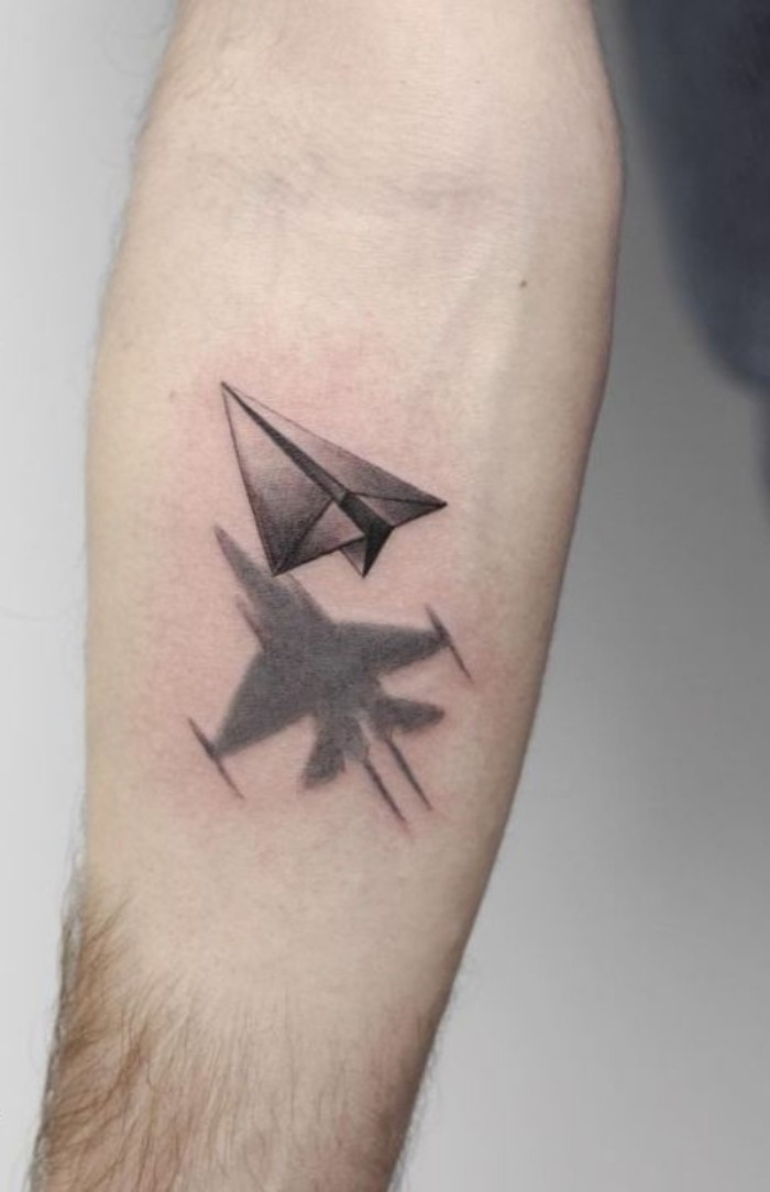 paper plane in grey, with a black outline, forearm tattoos, underneath it is a grey shadow, shaped a real plane