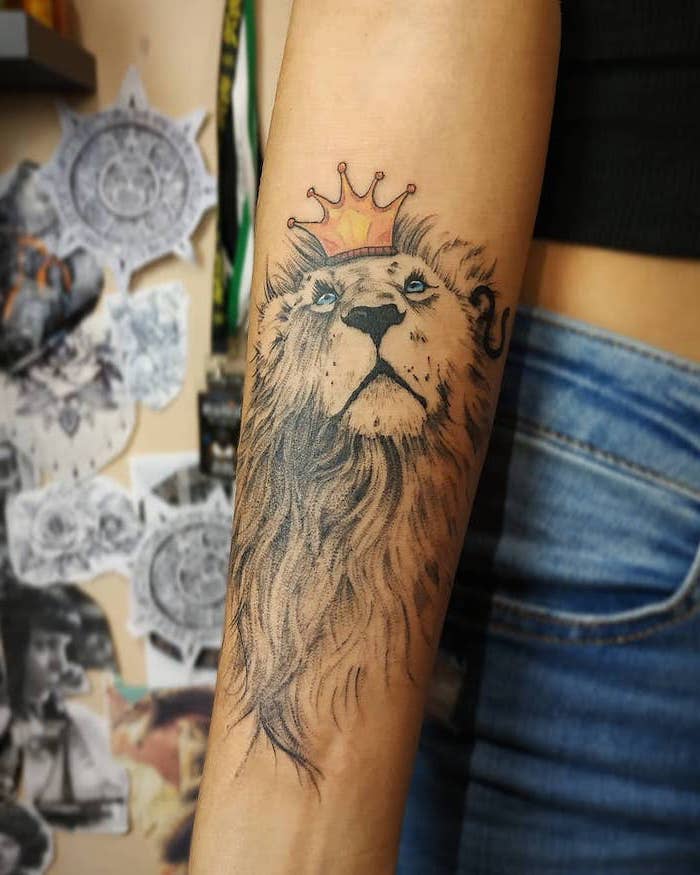 lion with long mane looking up, wearing a crown on its head, lion forearm tattoo, on woman wearing jeans and black top