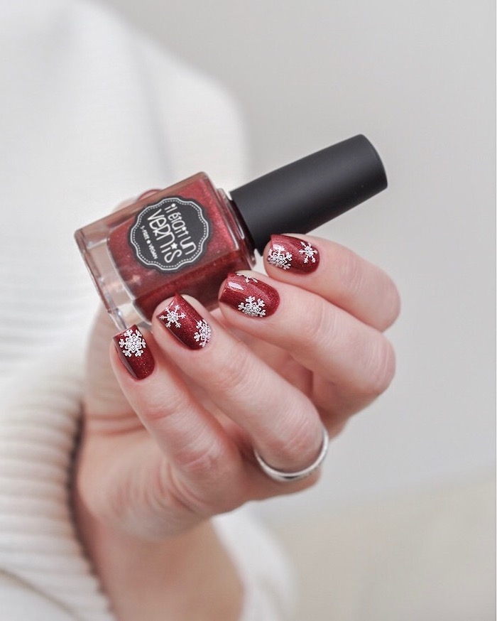 hand holding red nail polish bottle, short square nails with red nail polish, snowflakes decorations on each nail, winter nails