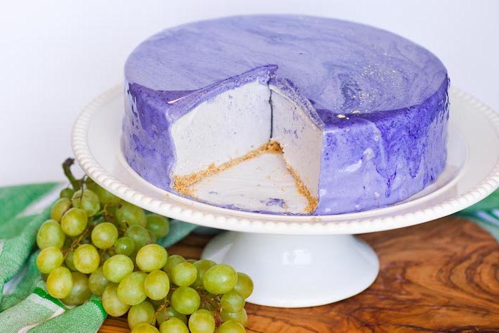 two tier cake, one tier cheesecake, covered with purple and white marble glaze, placed on white cake stand, grapes on the side